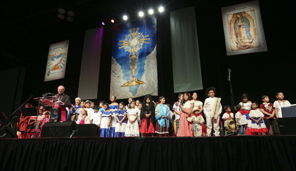 Wearing the traditional dress of their respective family's native country, children line the stage in the Spanish track after paying homage to Our Lady of Guadalupe with flowers. Bishop Joel M. Konzen, SM, left, called the children up by country. Photo By Michael Alexander