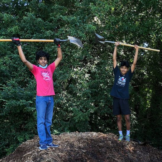 Two young male volunteers posting with shovels raised over their heads, standing on top of a large mound of mulch.