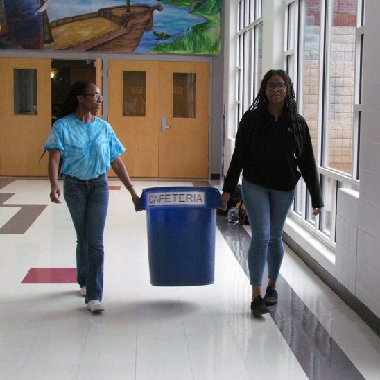 Two female students at Our Lady of Mercy Catholic High School (now St. Mary's Academy) carrying a cafeteria waste bin to properly dispose of it’s contents.